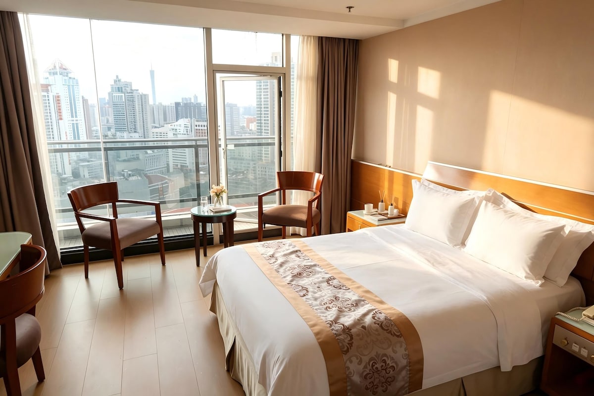 A hotel room with a large bed, two chairs, a small table, and a view of a cityscape through large windows.