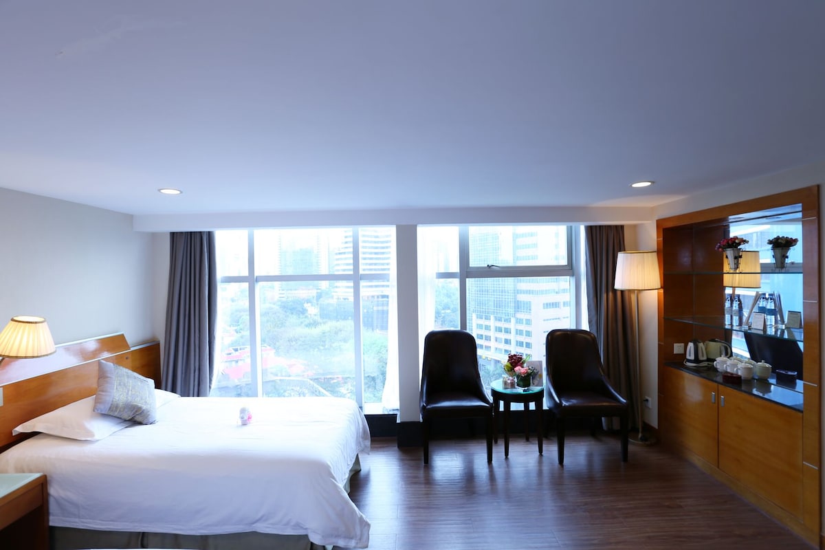 Une chambre d’hôtel avec un lit, deux chaises, une petite table, une fenêtre donnant sur la ville et un meuble en bois sur lequel sont posés divers objets.