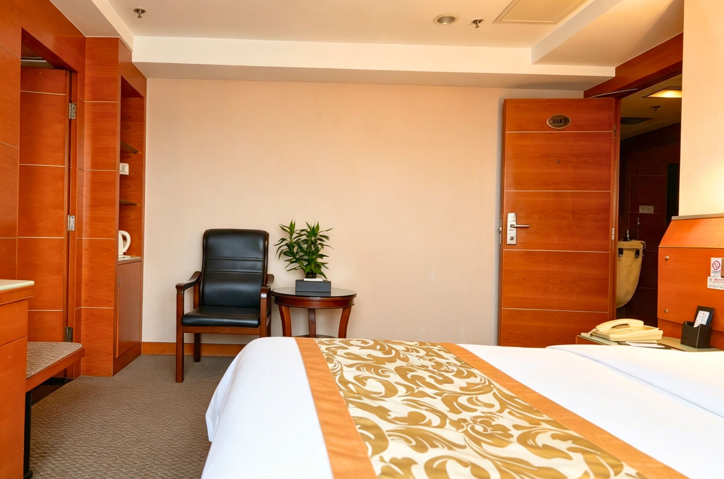 A hotel room with a bed, a desk, a chair, a small table with a plant, and a wardrobe.
