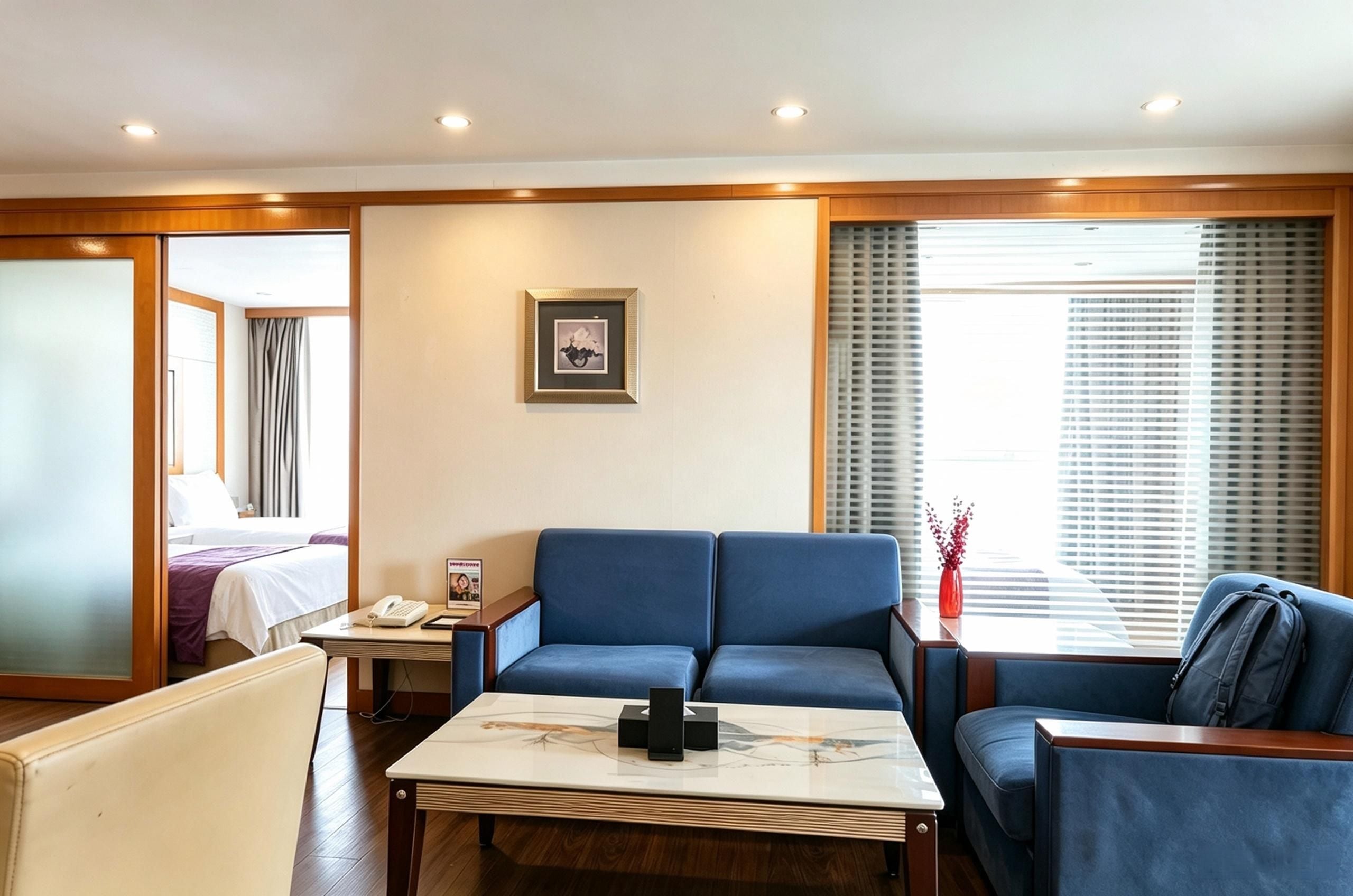 Une chambre d’hôtel avec un canapé bleu, une table basse, un lit avec une tête de lit blanche, une table de chevet avec un téléphone et un magazine, et une fenêtre avec des stores.