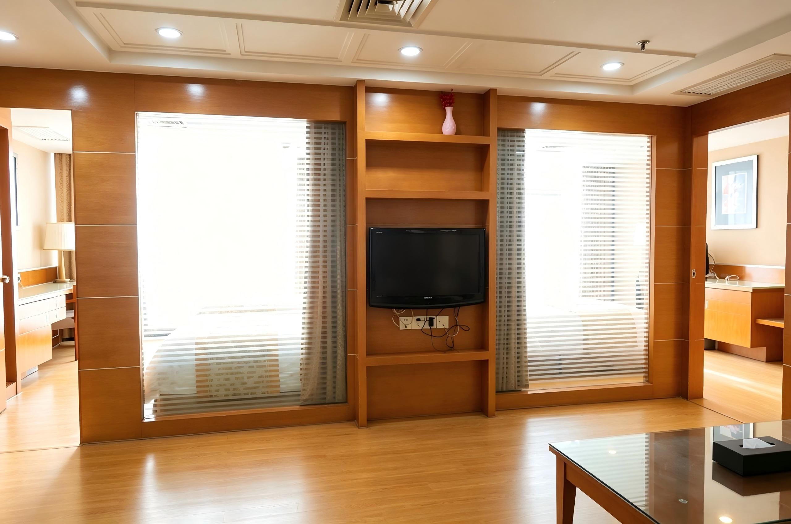 A modern living room with a flat-screen TV mounted on a wooden wall, wooden flooring, and a glass coffee table.