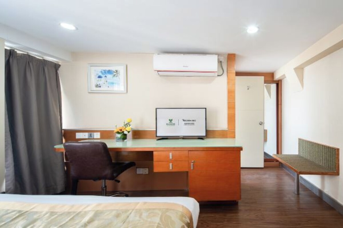 Une chambre d’hôtel avec un lit, un bureau avec une chaise, une petite table avec des fleurs, un climatiseur et un tableau encadré au mur.