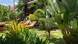 A tropical garden with a wooden staircase leading to a building, surrounded by lush greenery and palm trees.