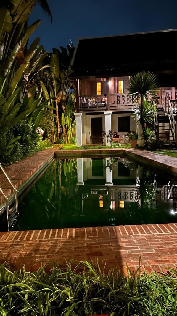 Una piscina con cubierta de madera, rodeada de abundante vegetación y un edificio de madera con balcón.
