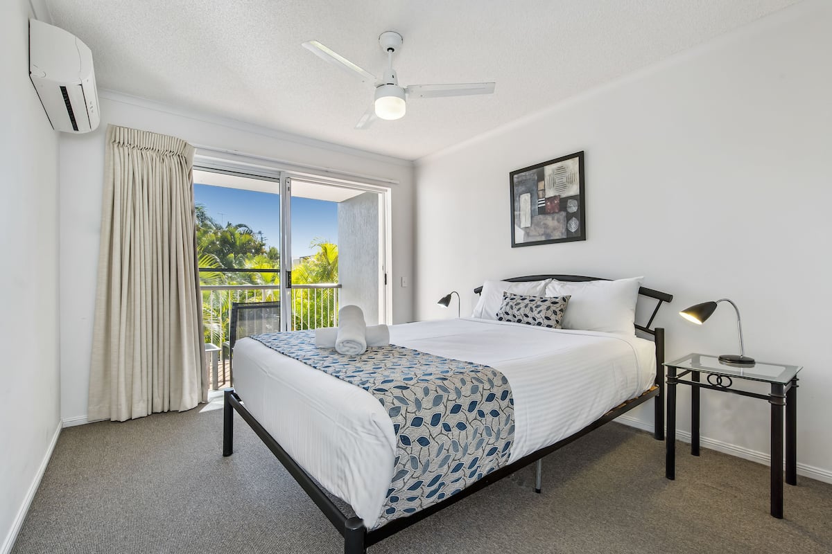 A hotel room with a bed, a ceiling fan, a window with curtains, a nightstand with a lamp, and a framed picture on the wall.
