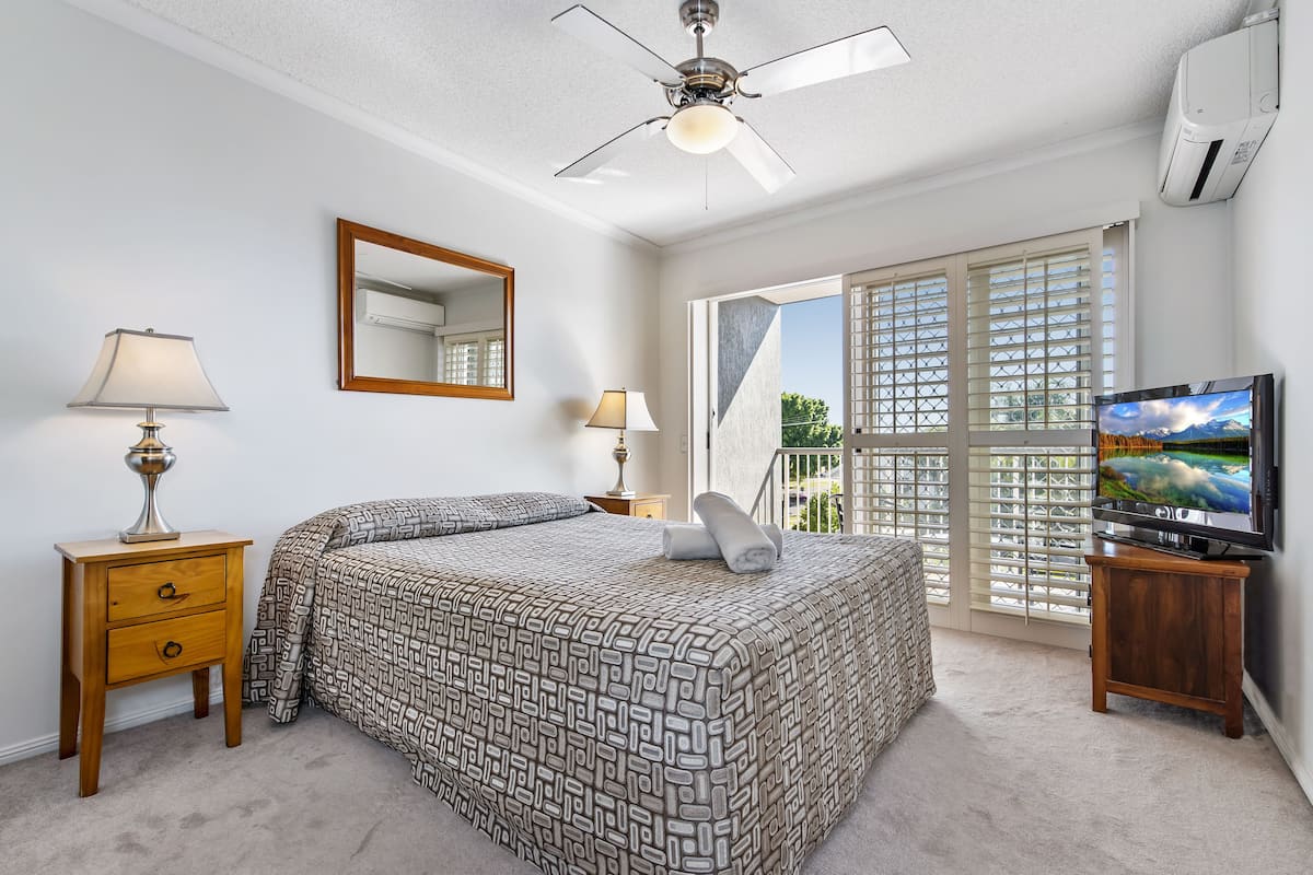 A bedroom with a bed, a nightstand with a lamp, a television, and a ceiling fan.