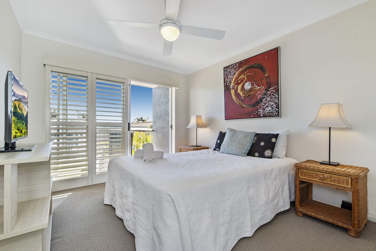 A bedroom with a bed, a desk, a TV, a ceiling fan, a painting, and a nightstand with lamps.