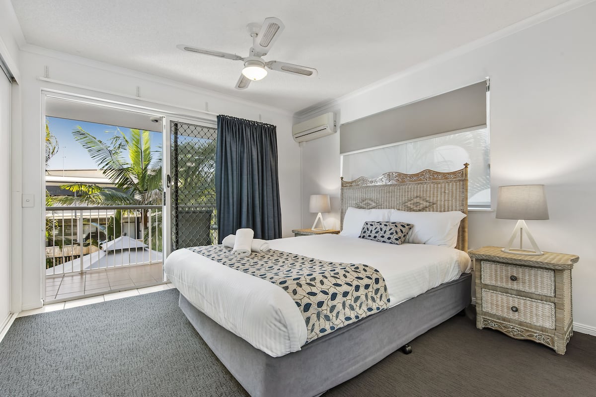 A hotel room with a large bed, a ceiling fan, a nightstand with a lamp, and a view of the outdoors.