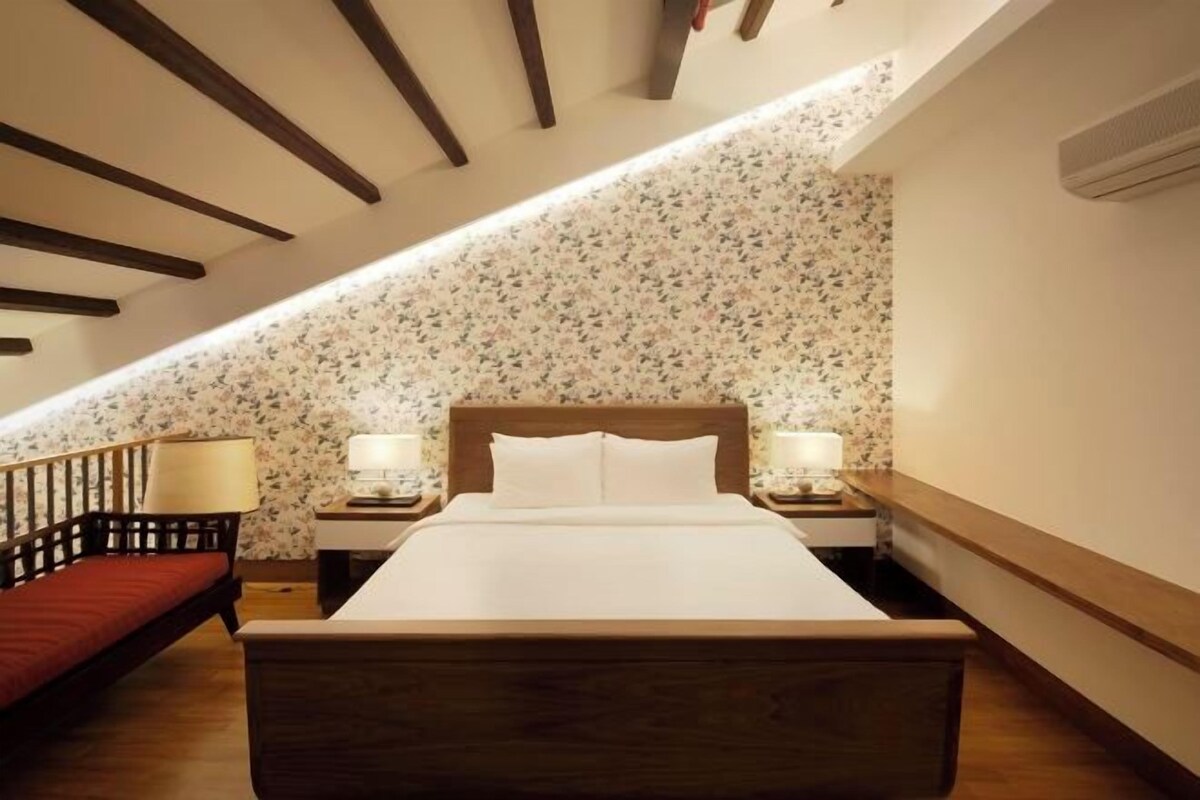 A bedroom with a wooden bed, two bedside tables with lamps, a red sofa, and a sloped ceiling with exposed beams.