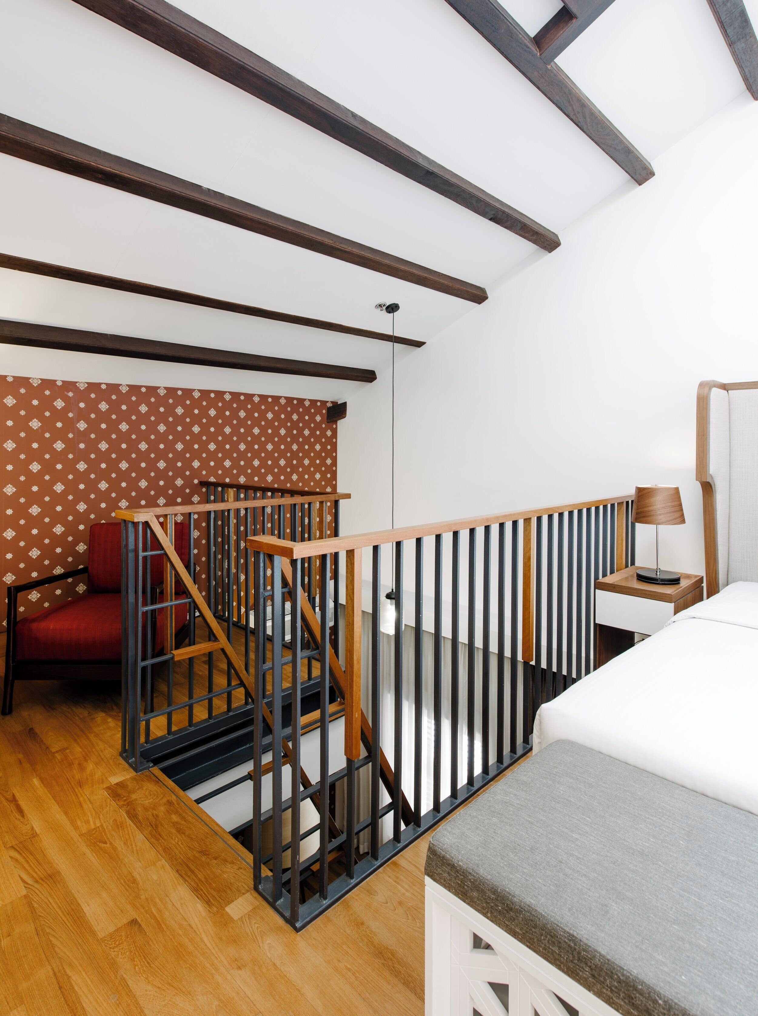 Una habitación con escalera de madera, cama, mesita de noche, lámpara y una silla.