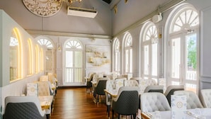 A restaurant with a long wooden floor, white walls, and arched windows. Tables are set with white tablecloths and silverware.