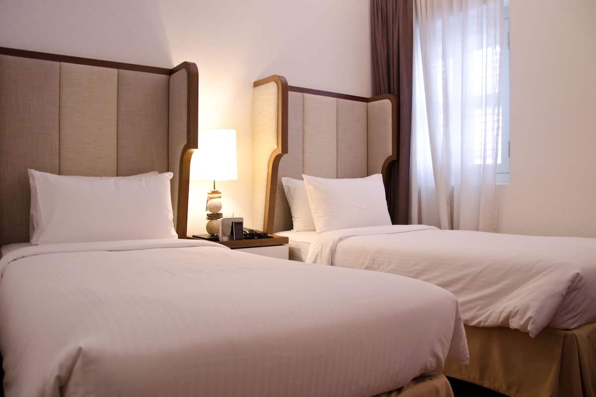 A hotel room with two beds, a nightstand with a lamp, and a window with curtains.