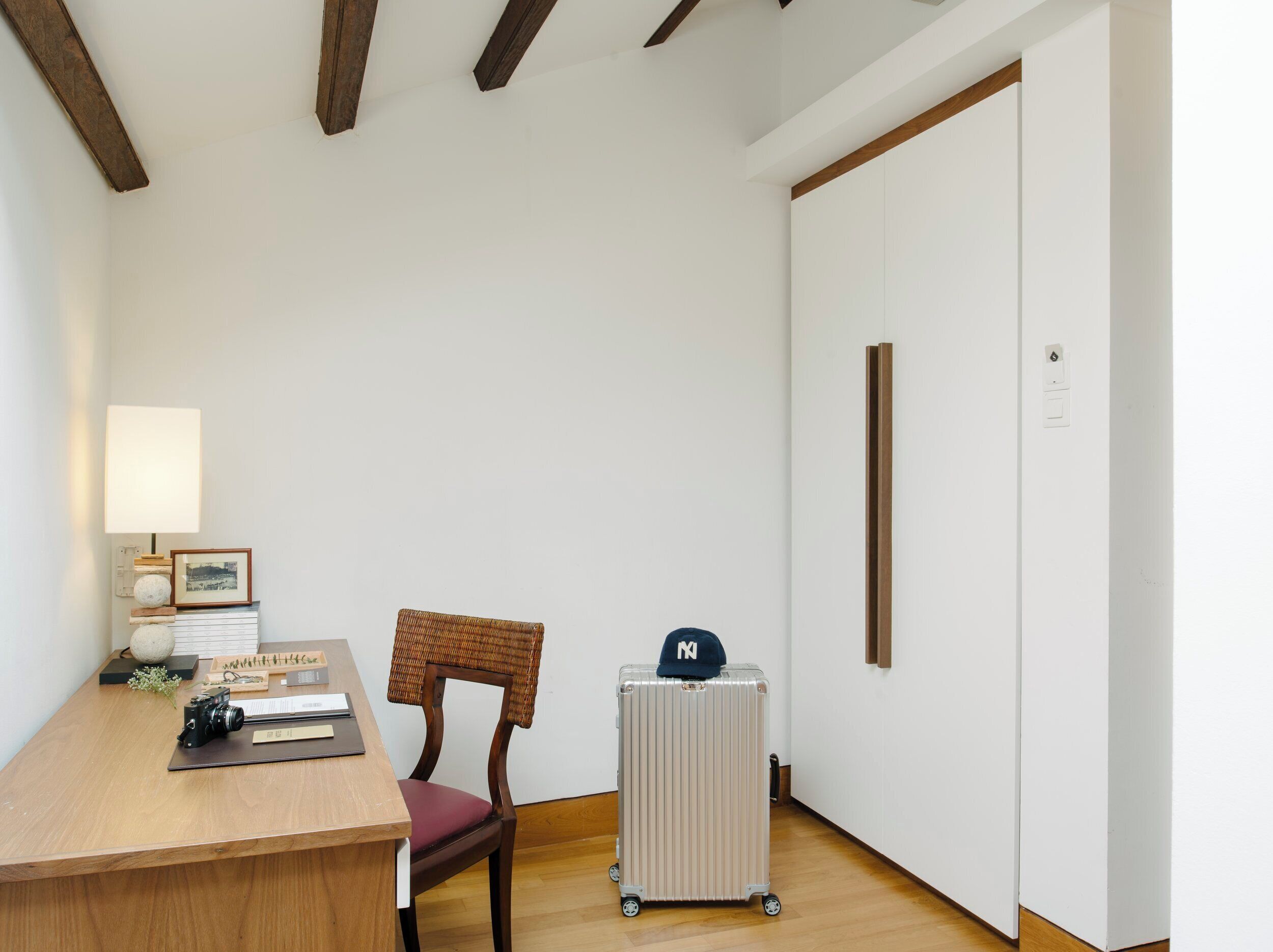 Quarto com escrivaninha de madeira, cadeira de vime, radiador e mala com tampa.