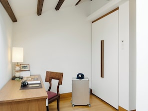 Une pièce avec un bureau en bois, une chaise en osier, un radiateur et une valise avec une casquette.
