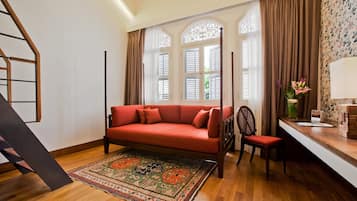 A room with a red sofa, a wooden chair, a patterned rug, and a desk with a lamp and a vase of flowers.