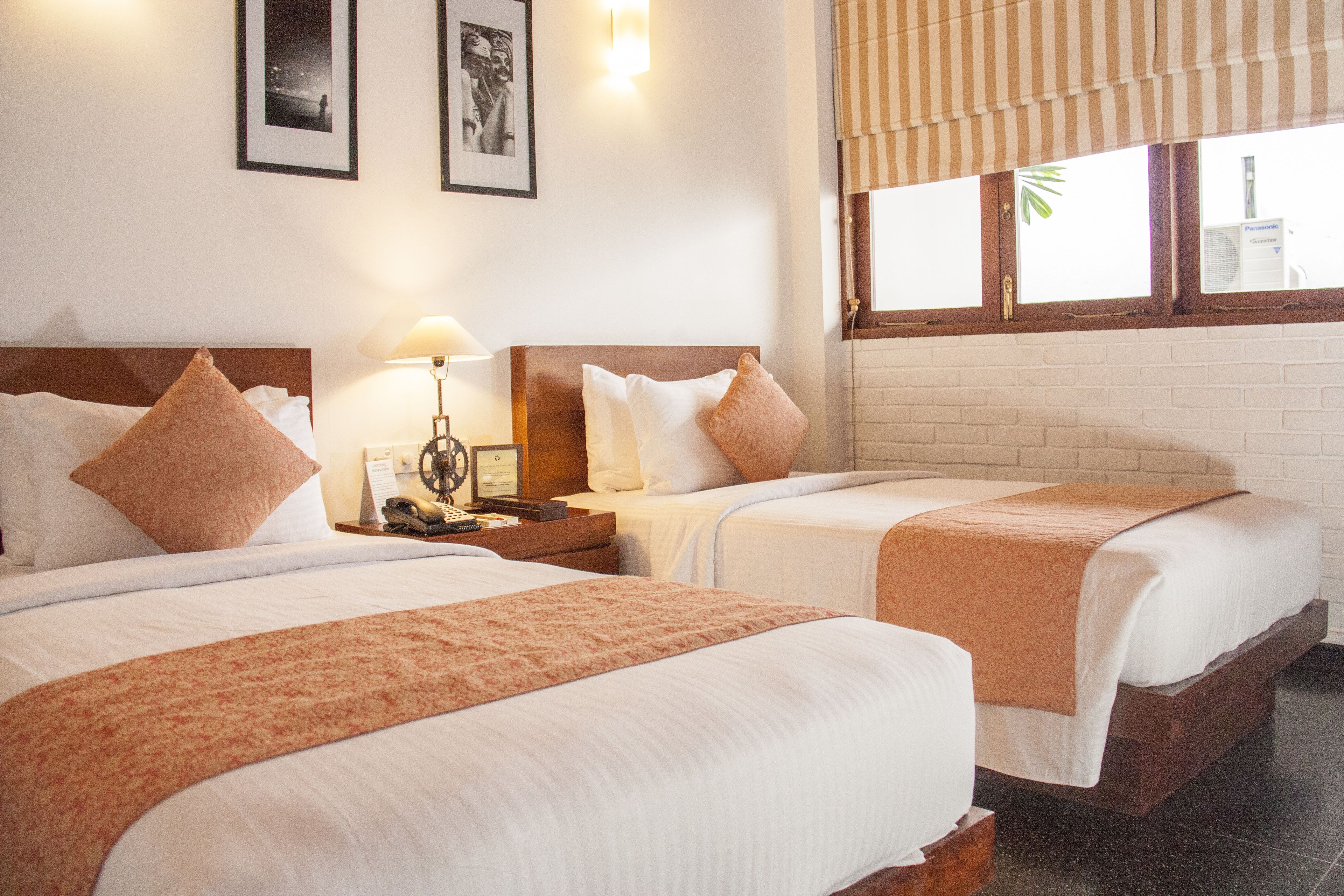 Habitación de hotel con dos camas, una mesita de noche con lámpara, cuadros en la pared y una ventana con vistas al exterior.