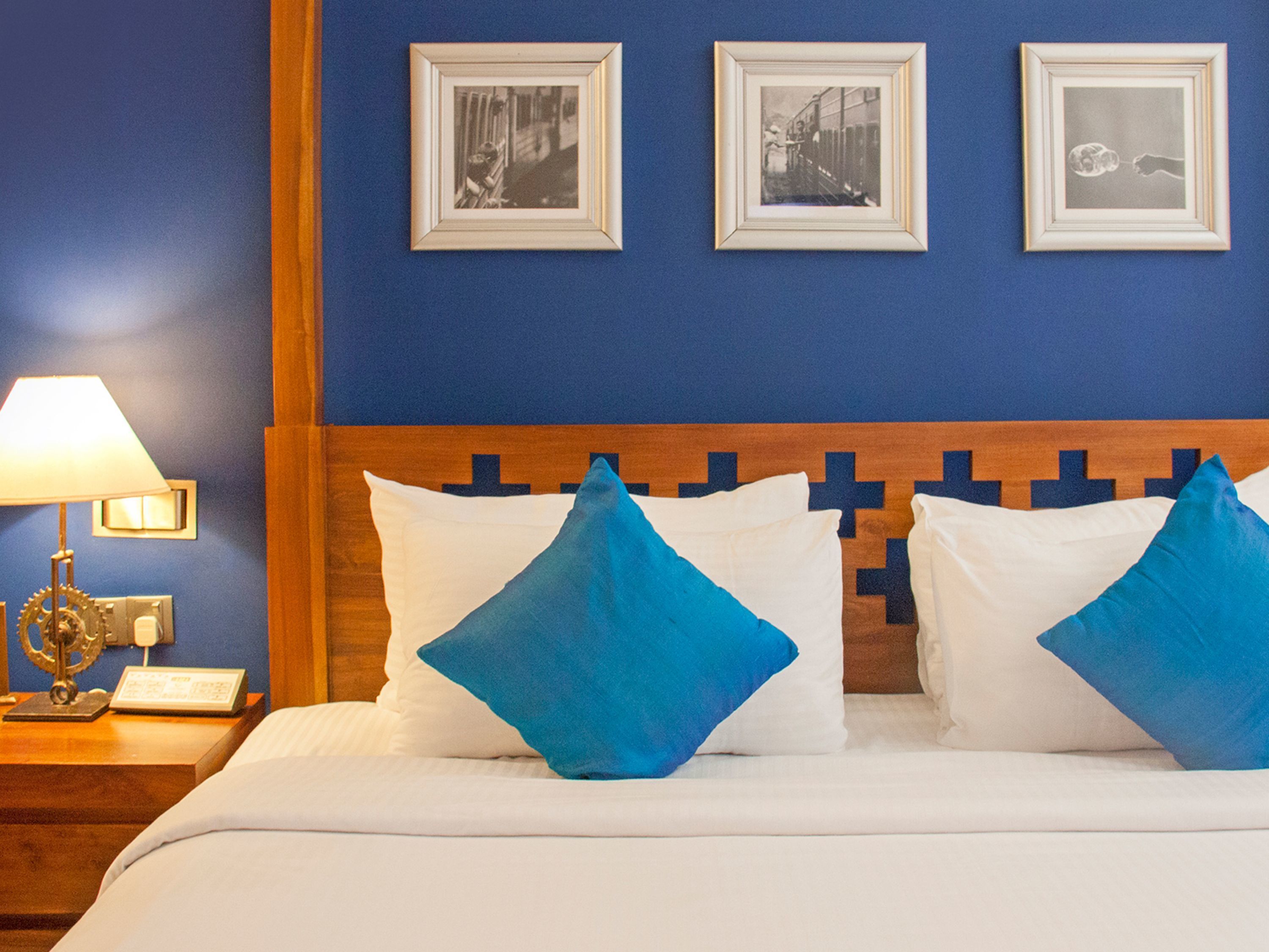 A hotel room with a bed, blue pillows, a wooden headboard, and three framed pictures on the wall.