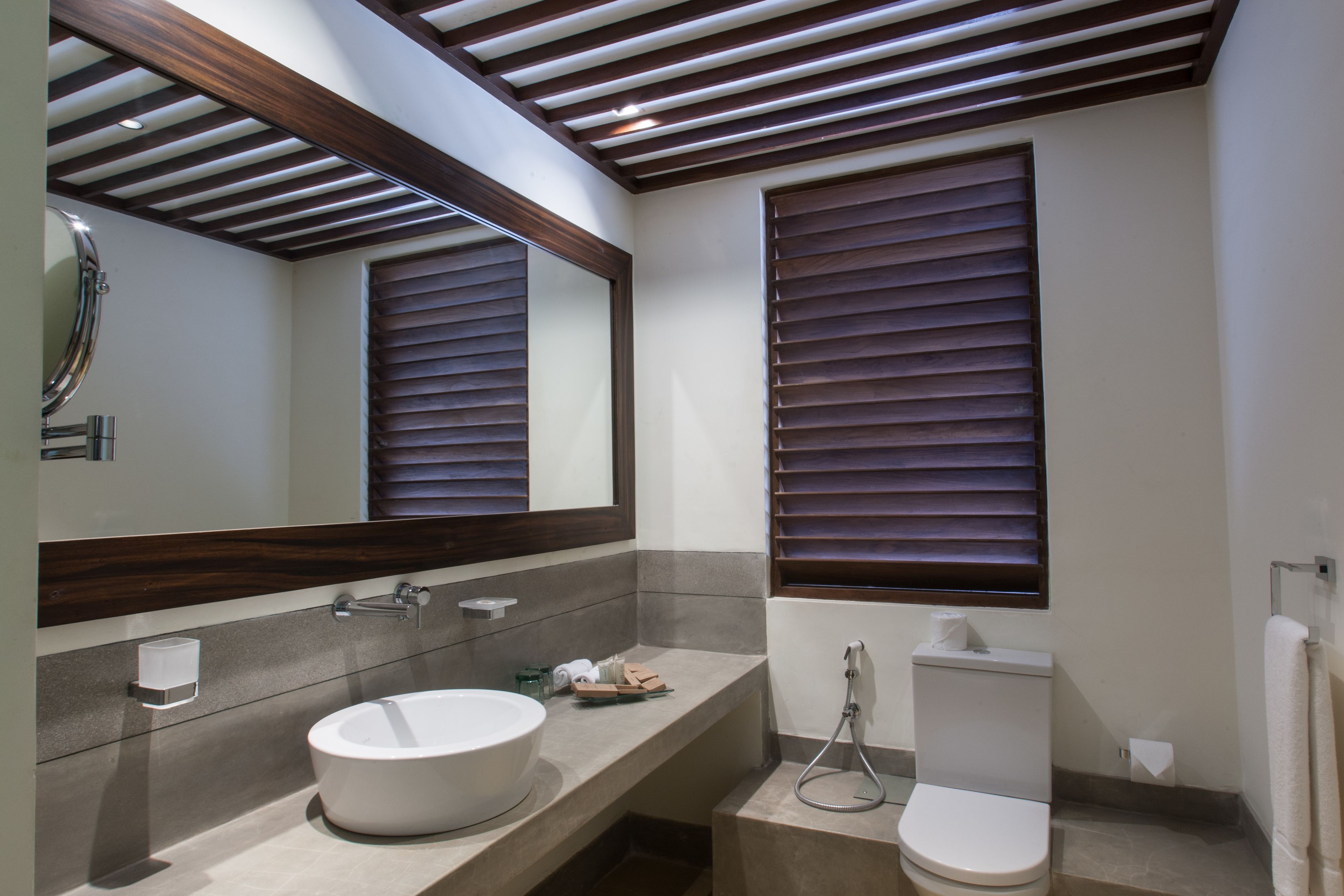 A modern bathroom with a large mirror, a round sink, a toilet, and a window with blinds.