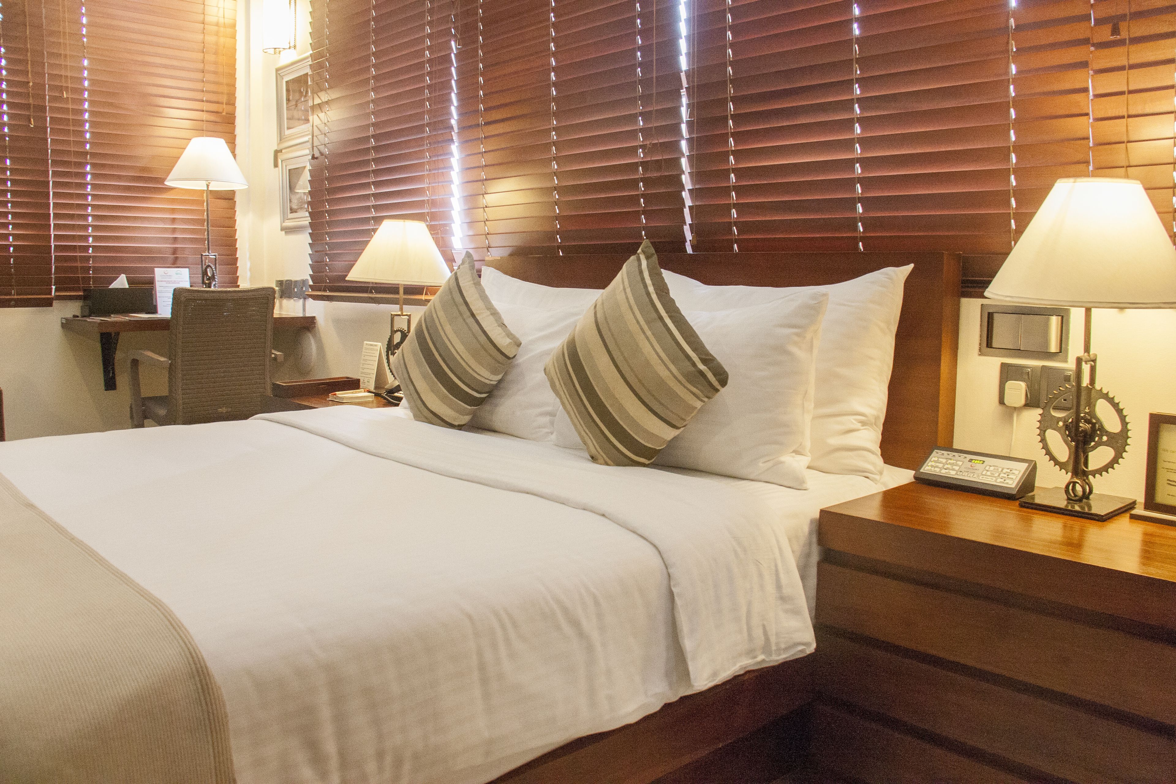 A hotel room with a large bed, two bedside lamps, a desk with a chair, and wooden blinds.