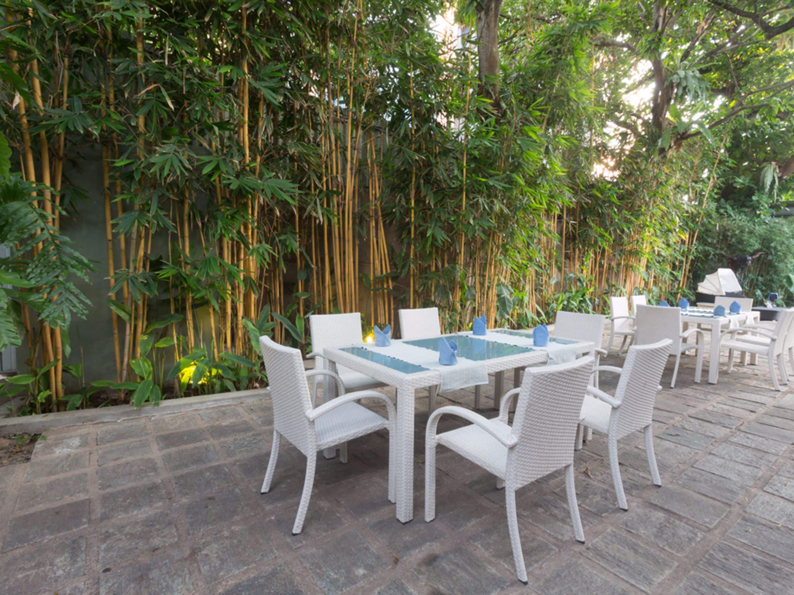 An outdoor dining area with white wicker furniture and a stone floor, surrounded by bamboo plants.