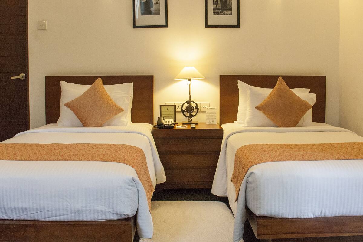 Camera d'albergo con due letti singoli, ciascuno con cuscino e coperta. Tra i letti, un comodino con una lampada e una cornice con un'immagine.