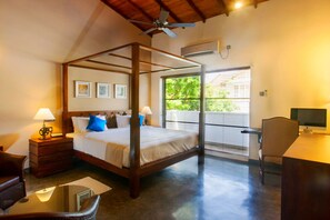 A four-poster bed with a wooden frame, two bedside tables with lamps, a desk with a computer, and a large window with blinds.