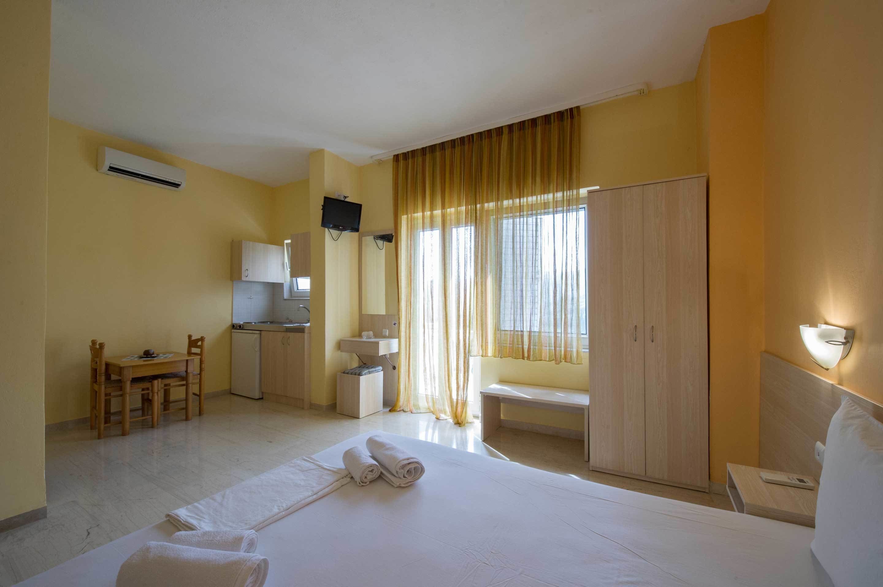 A hotel room with a bed, a desk, a chair, a small kitchenette, a wardrobe, and a window with curtains.
