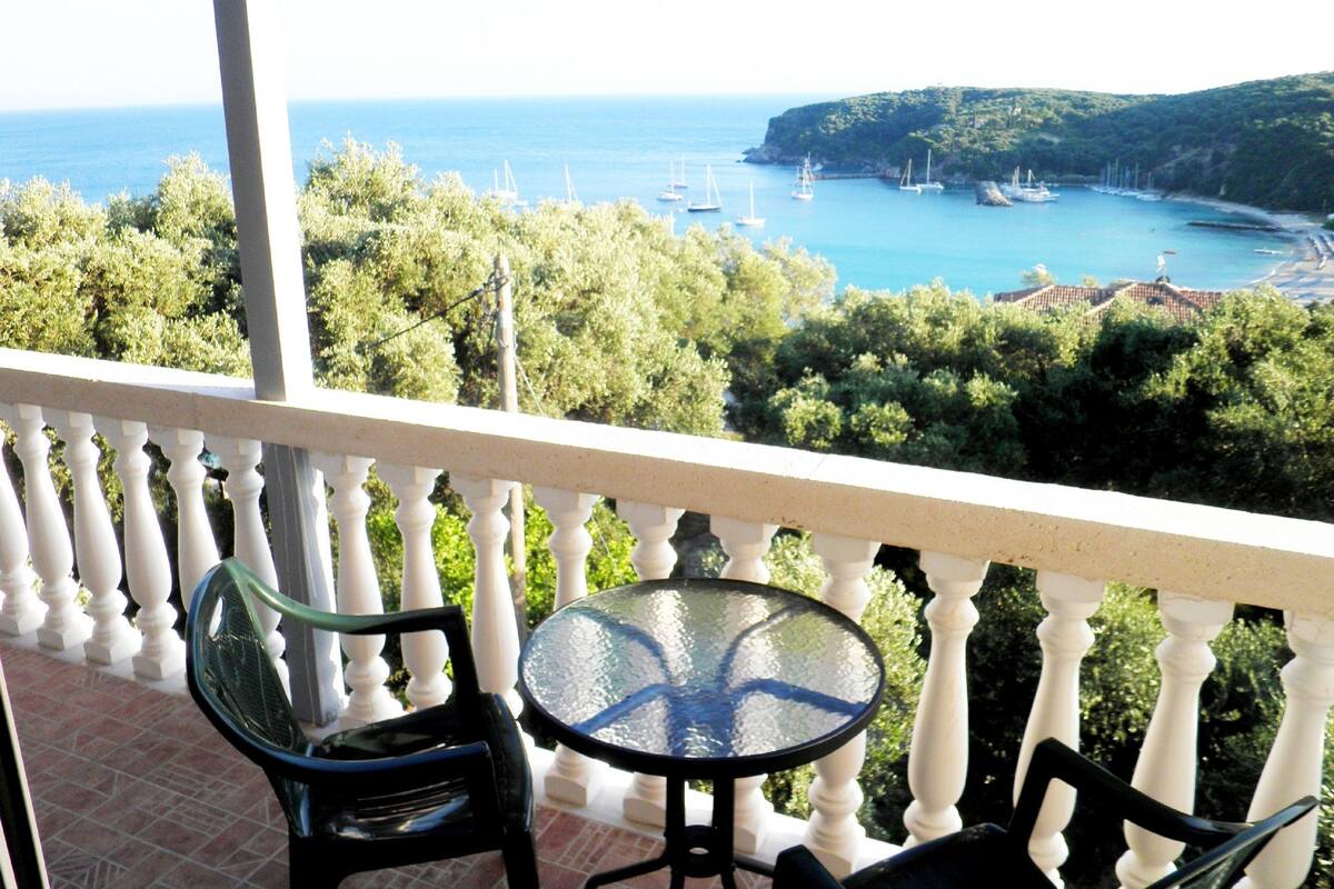 Ein Balkon mit Tisch und Stühlen mit Blick auf eine Küstenlandschaft mit Booten und viel Grün.