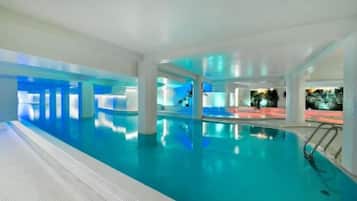 An indoor swimming pool with clear water, surrounded by white walls and columns, and a view of the night sky.