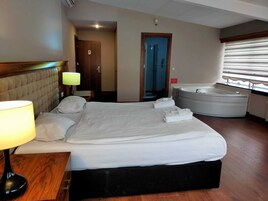 Habitación de hotel con una cama grande, una lámpara de noche, una bañera integrada y una ventana con persianas.