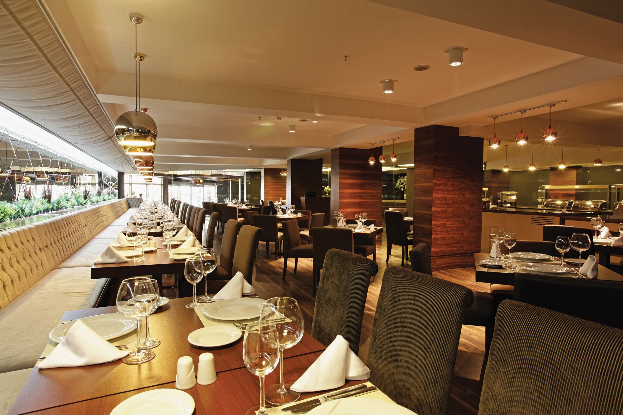Ein Restaurant mit langen Gemeinschaftstischen, separaten Sitzgelegenheiten und Blick auf Grün durch große Fenster.
