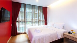 Une chambre d’hôtel avec un grand lit, un téléviseur à écran plat fixé au mur, une table de chevet avec un téléphone et un vase de fleurs, et une fenêtre avec des rideaux légers.