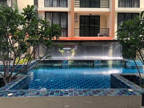 Una piscina con mosaicos azules, rodeada de plantas y un edificio con balcones.