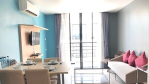 Une chambre d’hôtel moderne avec un coin repas, un canapé agrémenté de coussins roses et une grande fenêtre avec des rideaux.
