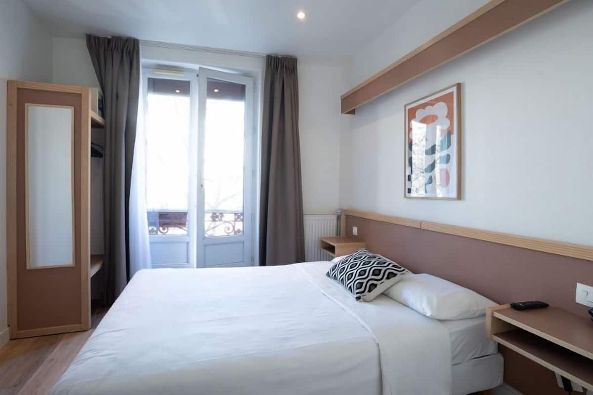 A hotel room with a bed, a nightstand, a painting, and a view of the outside.