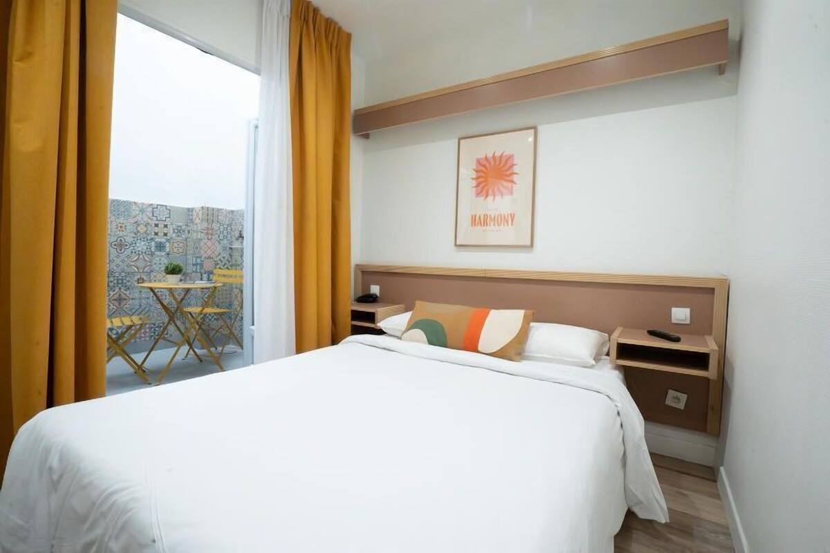 A hotel room with a bed, a nightstand, a framed picture on the wall, and a window with curtains.
