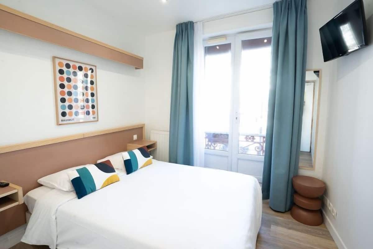 A hotel room with a bed, a bedside table, a mounted TV, and a window with curtains.