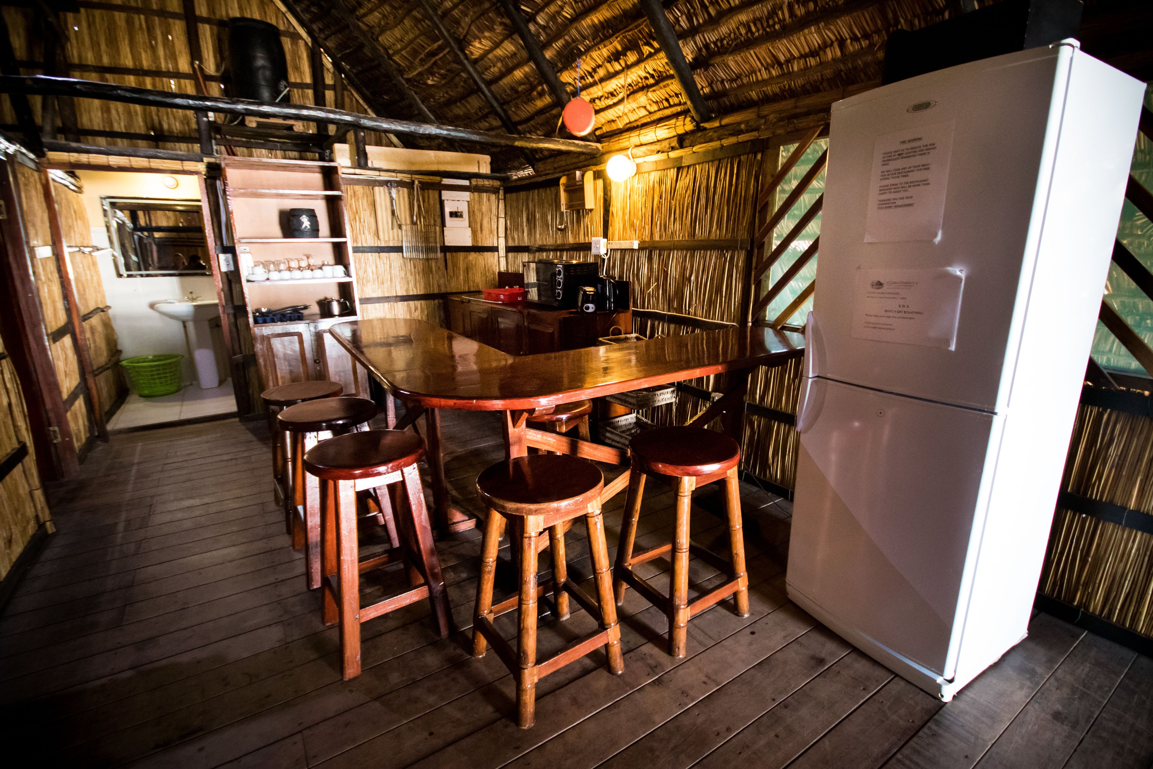 Uma cozinha rústica com bancos de madeira, uma mesa de madeira, uma geladeira branca e um telhado de palha.