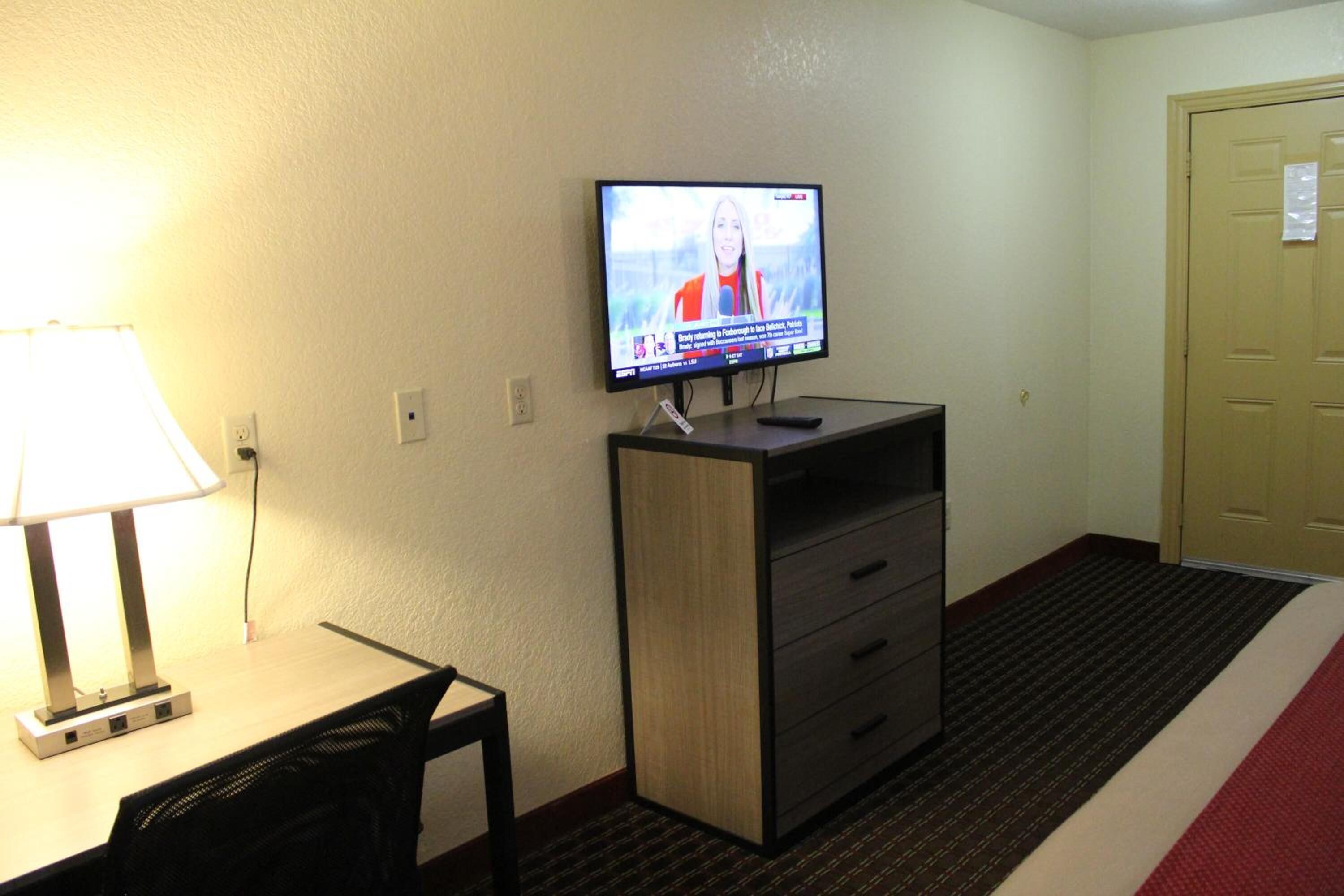 Hotelkamer met een televisie op een standaard, een bureau met een lamp, een stoel, een kledingkast en een vloerbedekking.