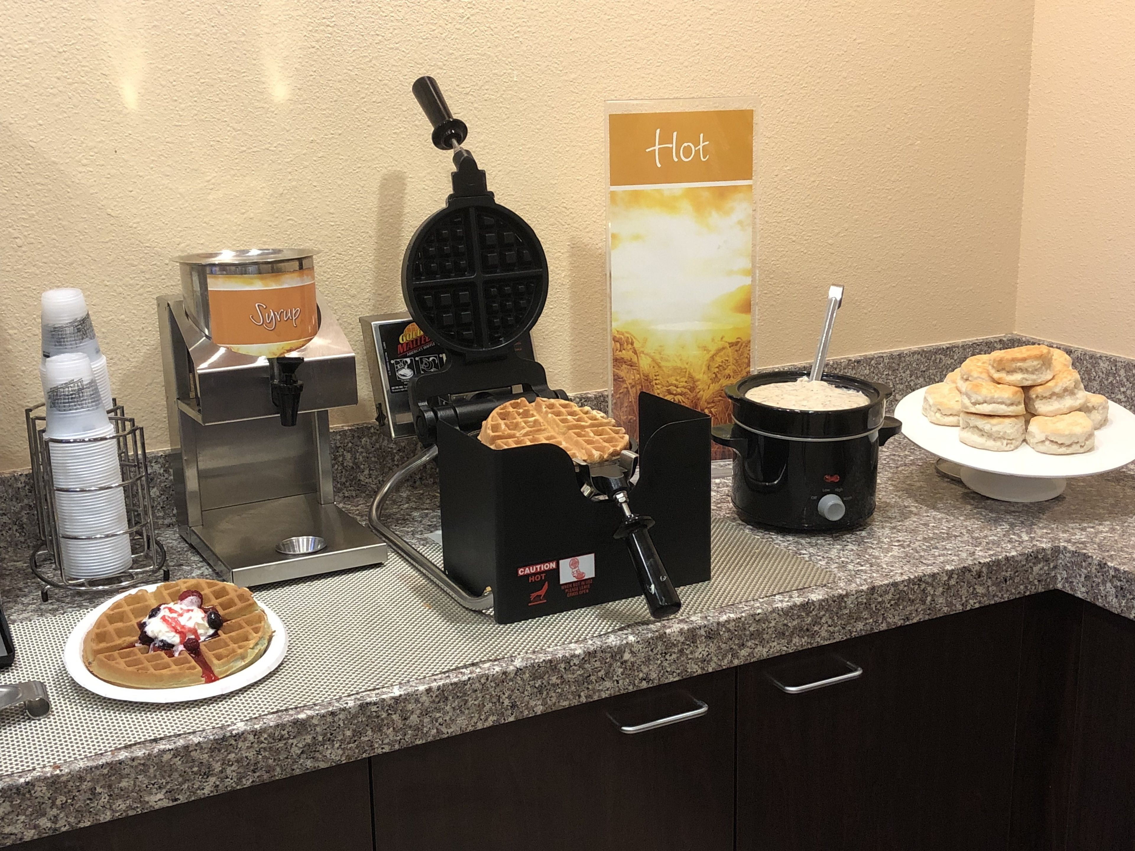 Uma estação de café da manhã com máquina de waffles, dosador de calda e estação de comidas quentes.