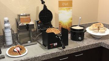 Uma estação de café da manhã com máquina de waffles, dosador de calda e estação de comidas quentes.