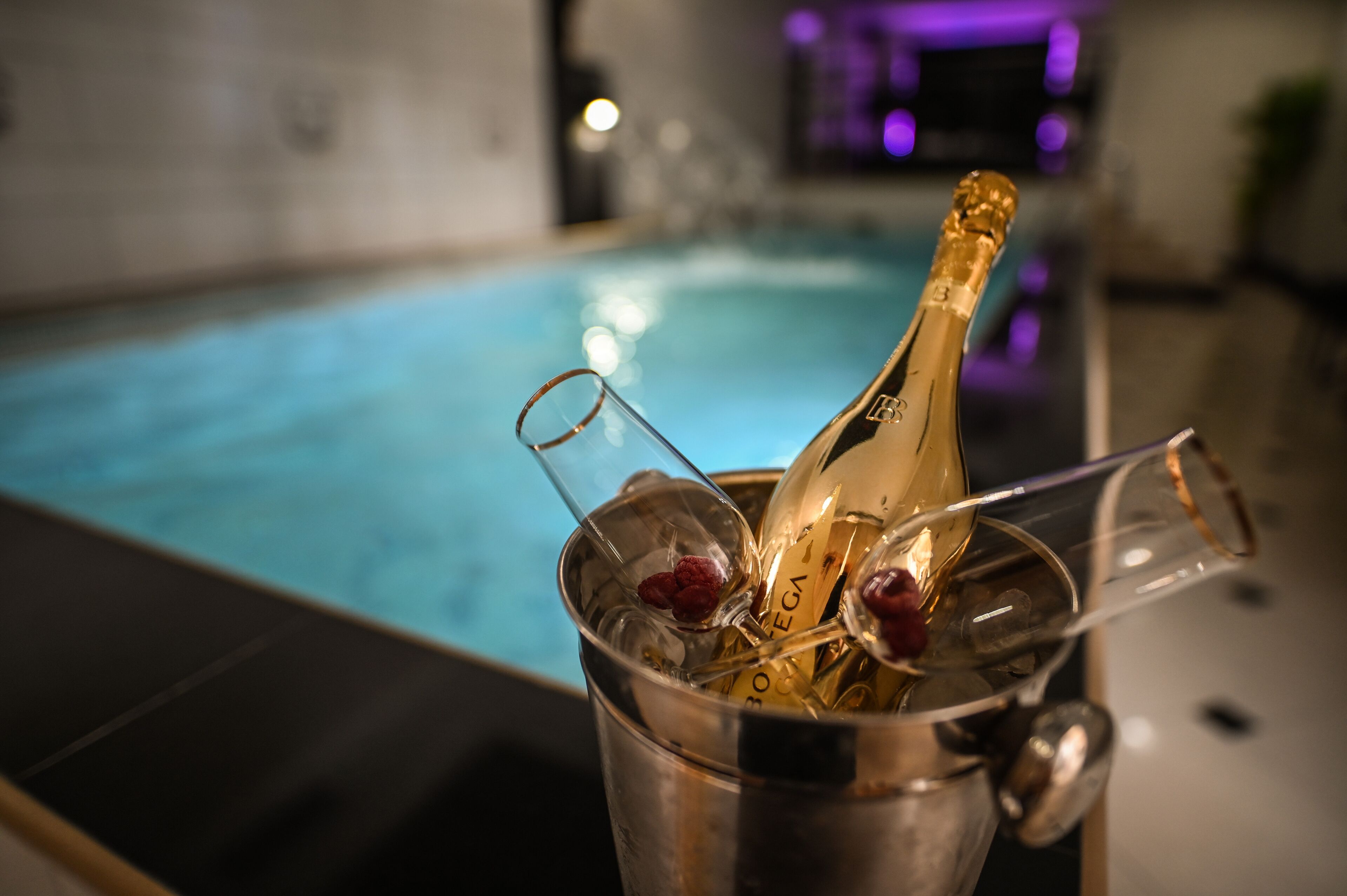 A champagne bottle in a metal bucket with glasses and a spoon, near a pool.