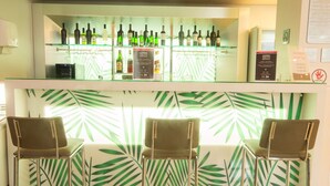 Una zona de bar con un estante de vidrio que exhibe varias botellas, dos sillas y una pared con diseño de hojas de palma.
