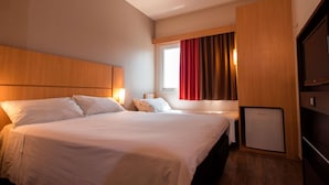 Habitación de hotel con dos camas, cabecero de madera, un televisor pequeño y una ventana con cortinas.