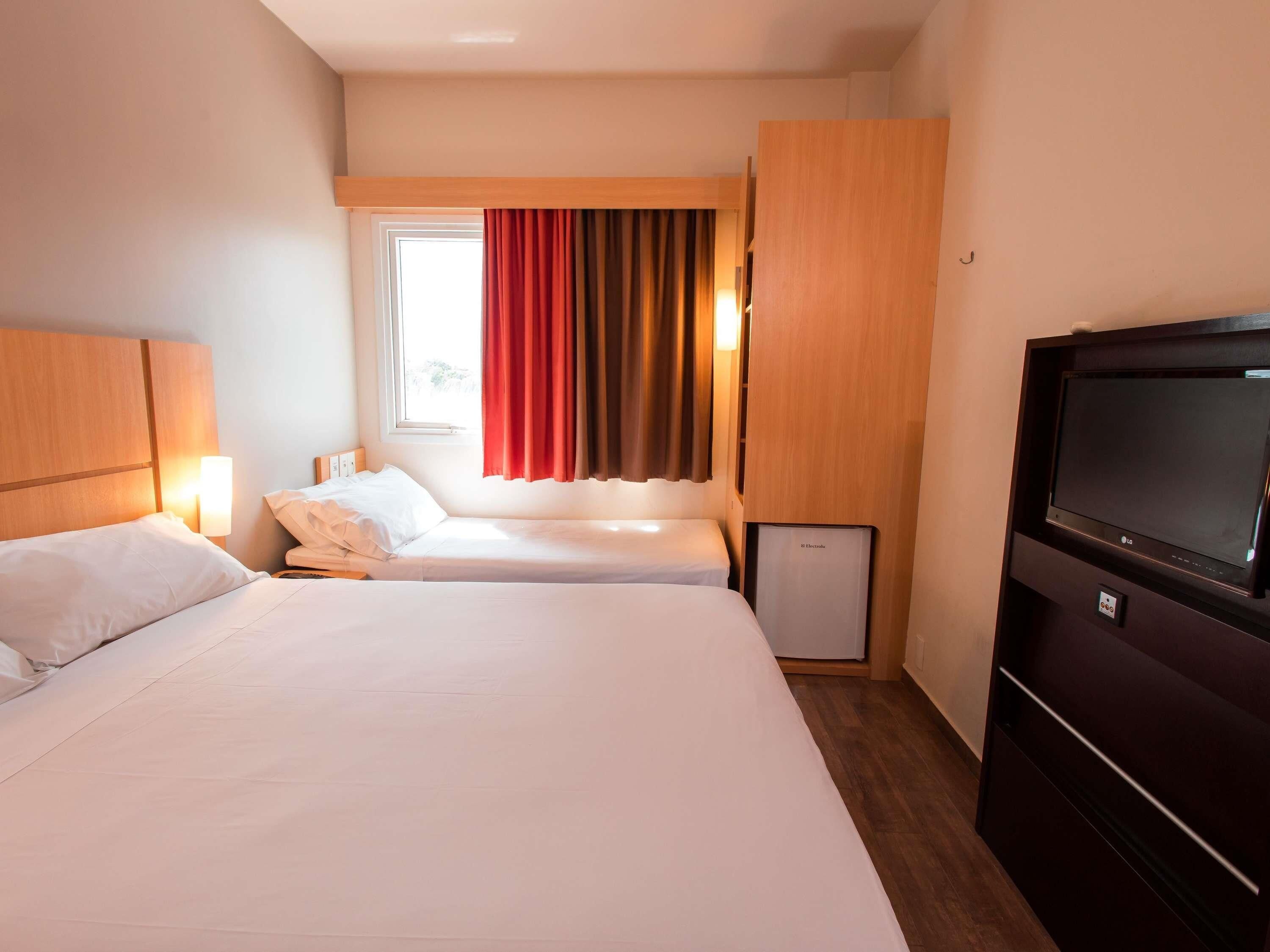 Camera d'albergo con un letto grande, un letto più piccolo, una TV e una finestra con tende.