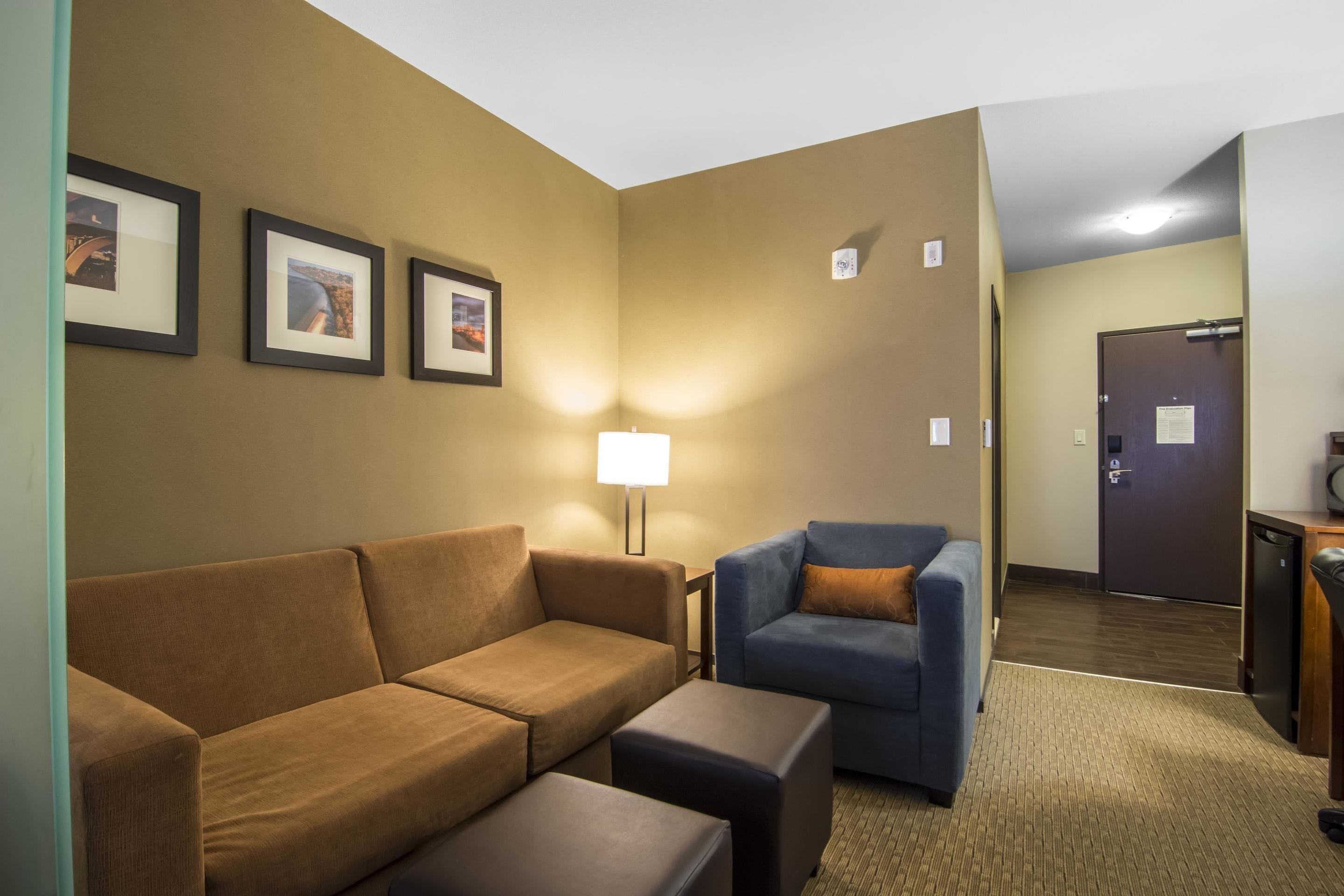 Habitación de hotel con un sofá marrón, un sillón azul y una mesita. En la pared hay cuadros enmarcados y una lámpara sobre la mesita.