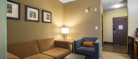 Habitación de hotel con un sofá marrón, un sillón azul y una mesita. En la pared hay cuadros enmarcados y una lámpara sobre la mesita.