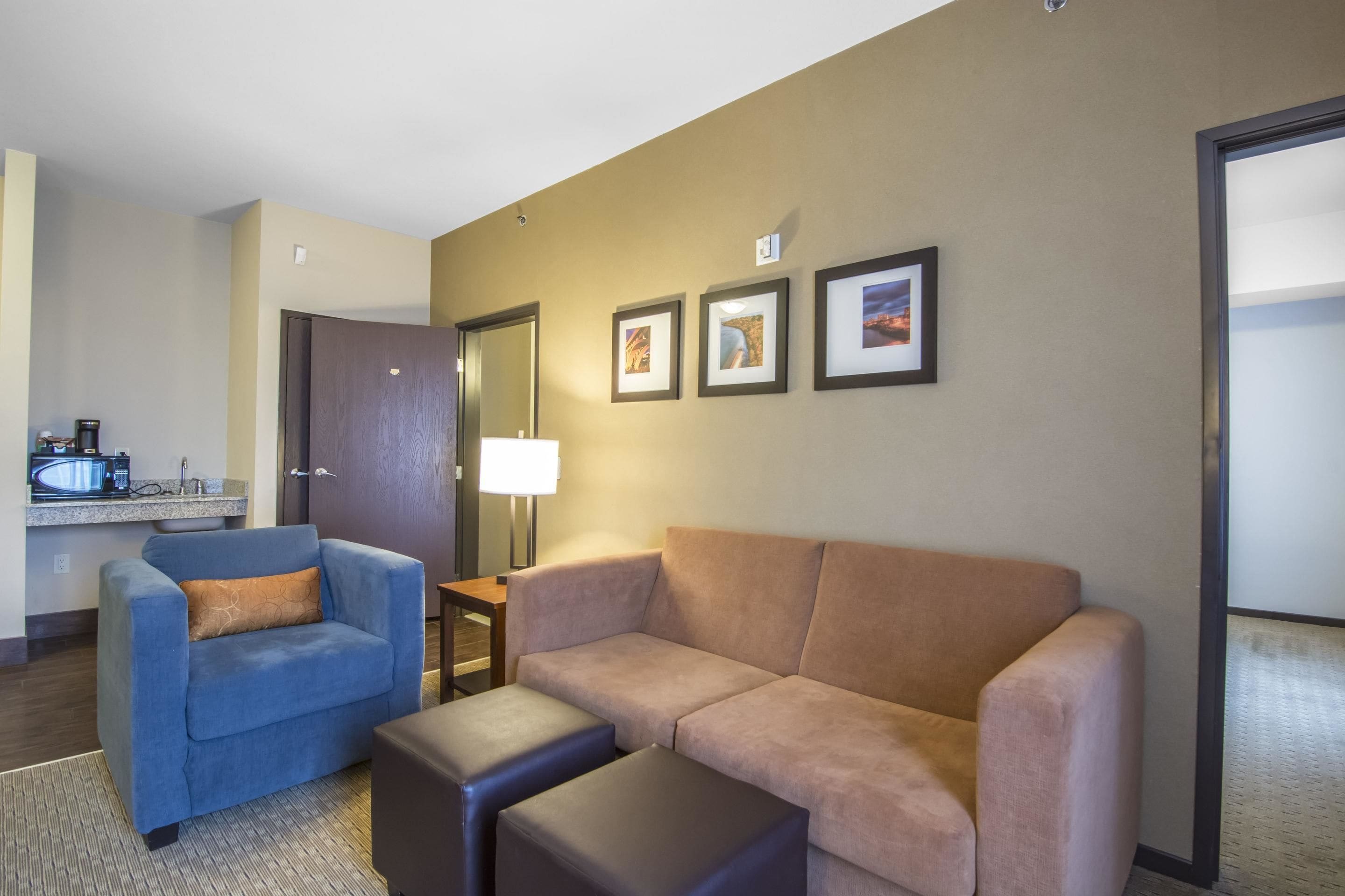 A hotel room with a sofa, armchair, and a small table with a lamp. There is a kitchenette with a microwave and coffee maker.