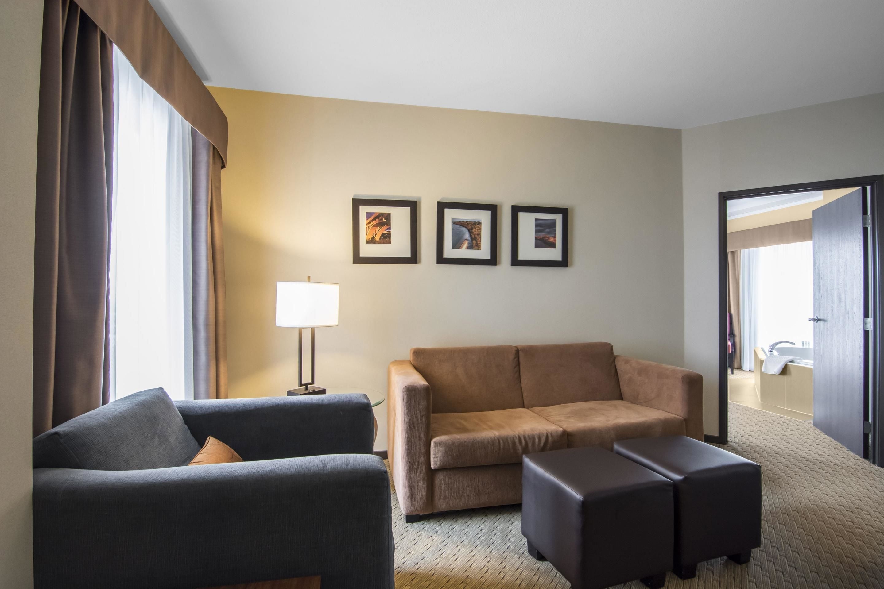 Ein Hotelzimmer mit einem dunkelgrauen Sofa, einem braunen Sofa und einem passenden Hocker. An der Wand hängen drei gerahmte Bilder, eine Lampe ist vorhanden und durch eine geöffnete Tür ist das Badezimmer sichtbar.