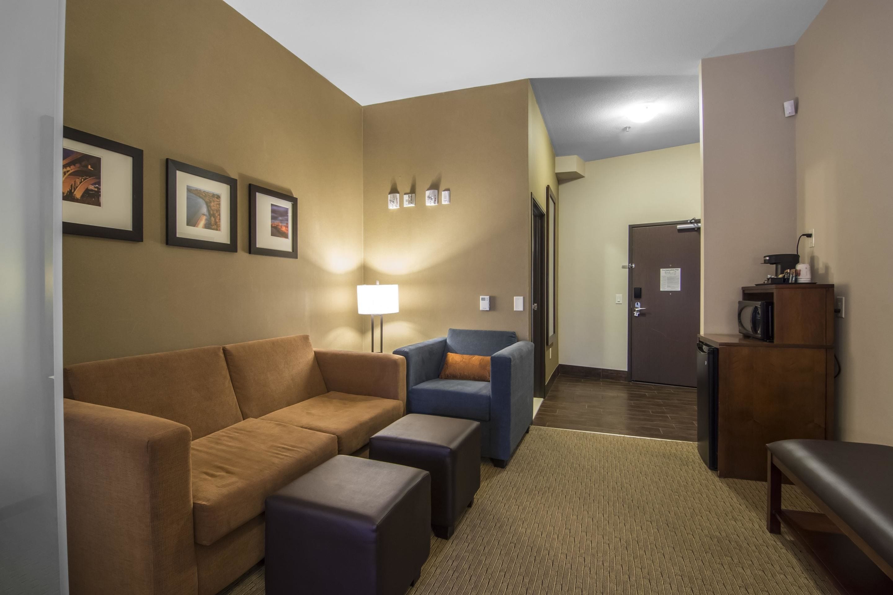 Habitación de hotel con sofá marrón, sillón azul, una mesita pequeña y un banco. Hay una lámpara, un refrigerador y un microondas sobre un armario de madera.